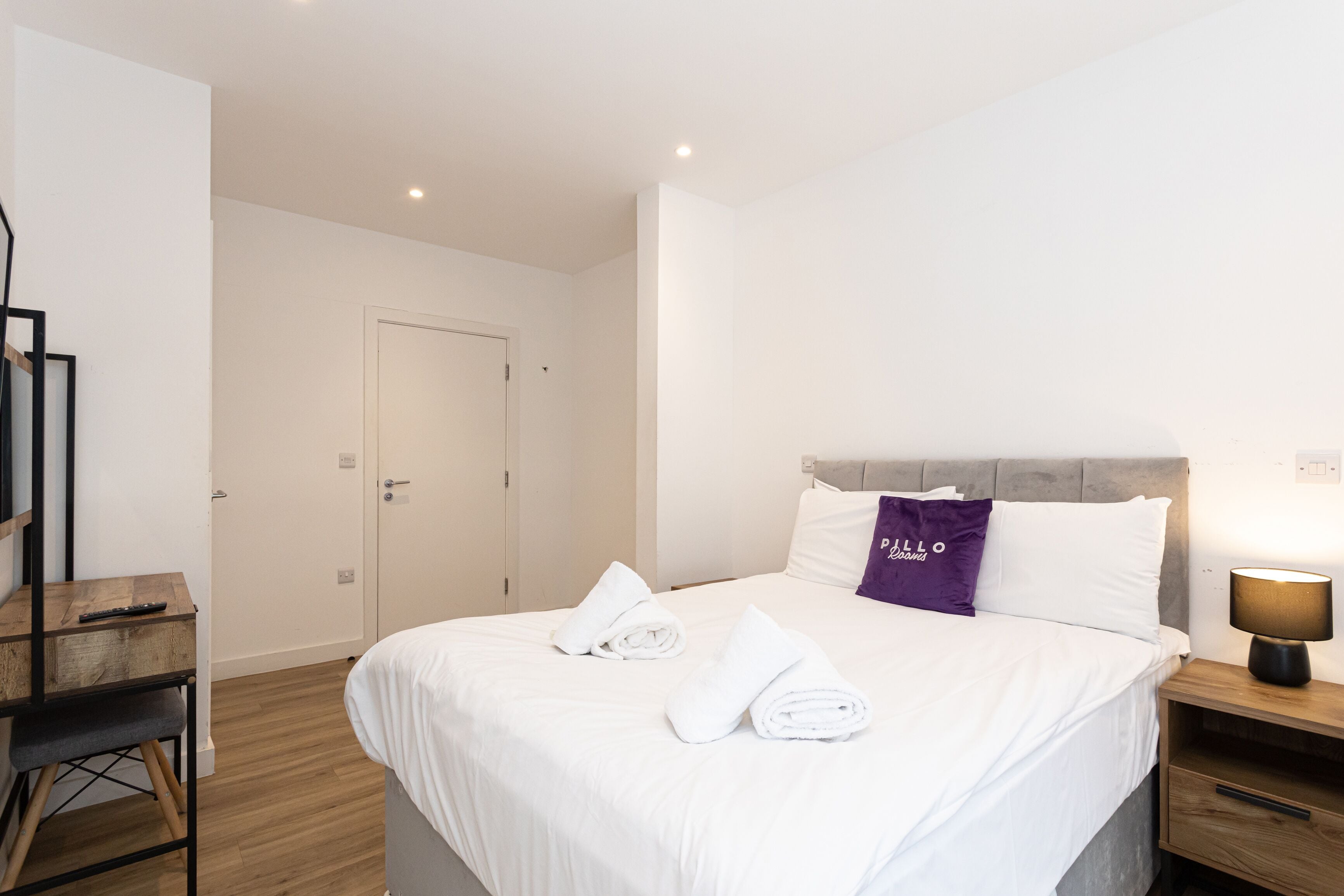 A neatly made bed with white linens and a purple pillow, a wooden nightstand with a lamp, and a wooden desk with a chair.