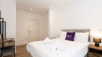 A neatly made bed with white linens and a purple pillow, a wooden nightstand with a lamp, and a wooden desk with a chair.