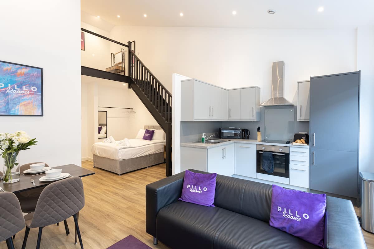A modern apartment with a dining area, a sofa with purple pillows, a kitchen, and a staircase.