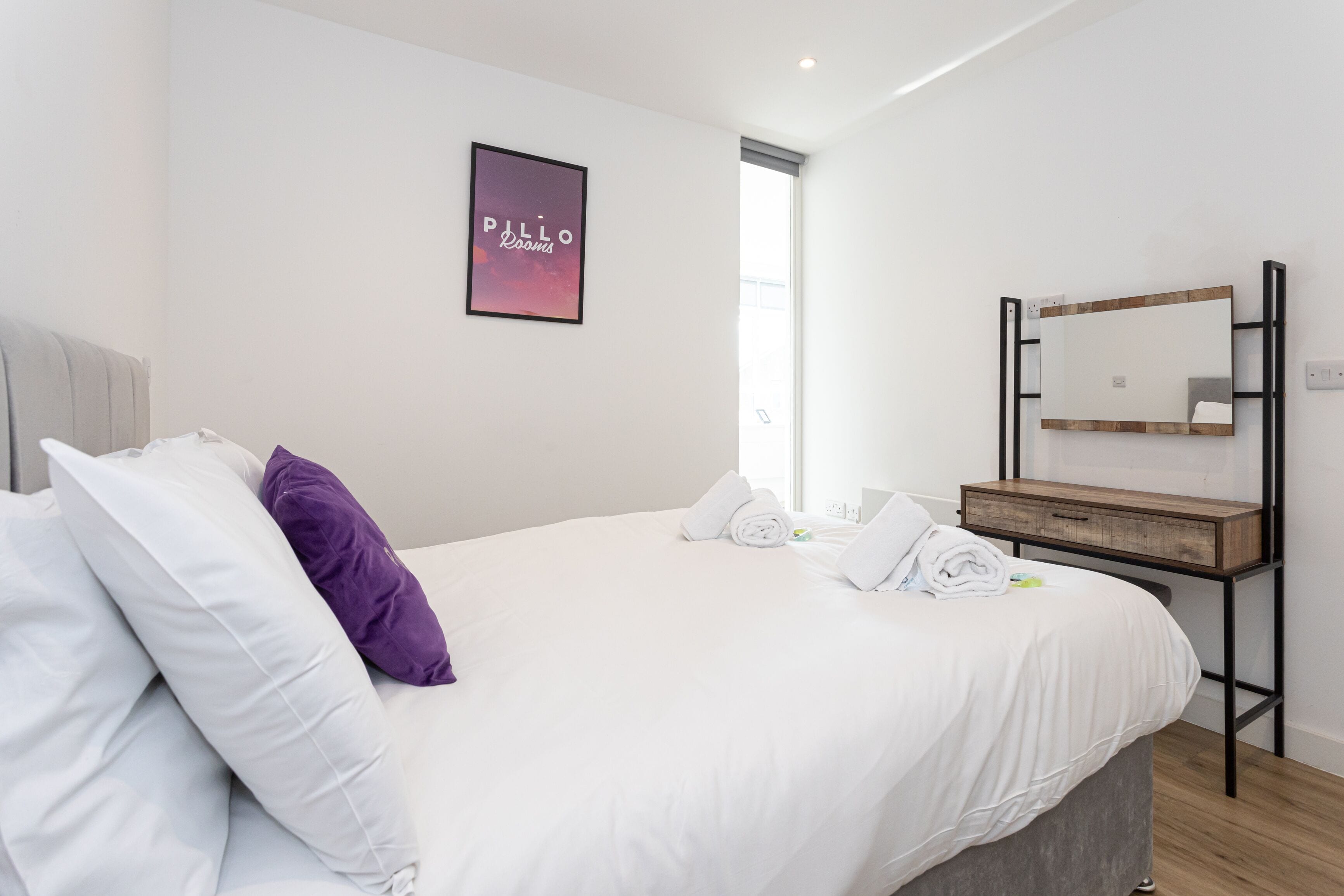 A neatly made bed with white and purple pillows, a framed picture on the wall, and a wooden bedside table with a mirror.