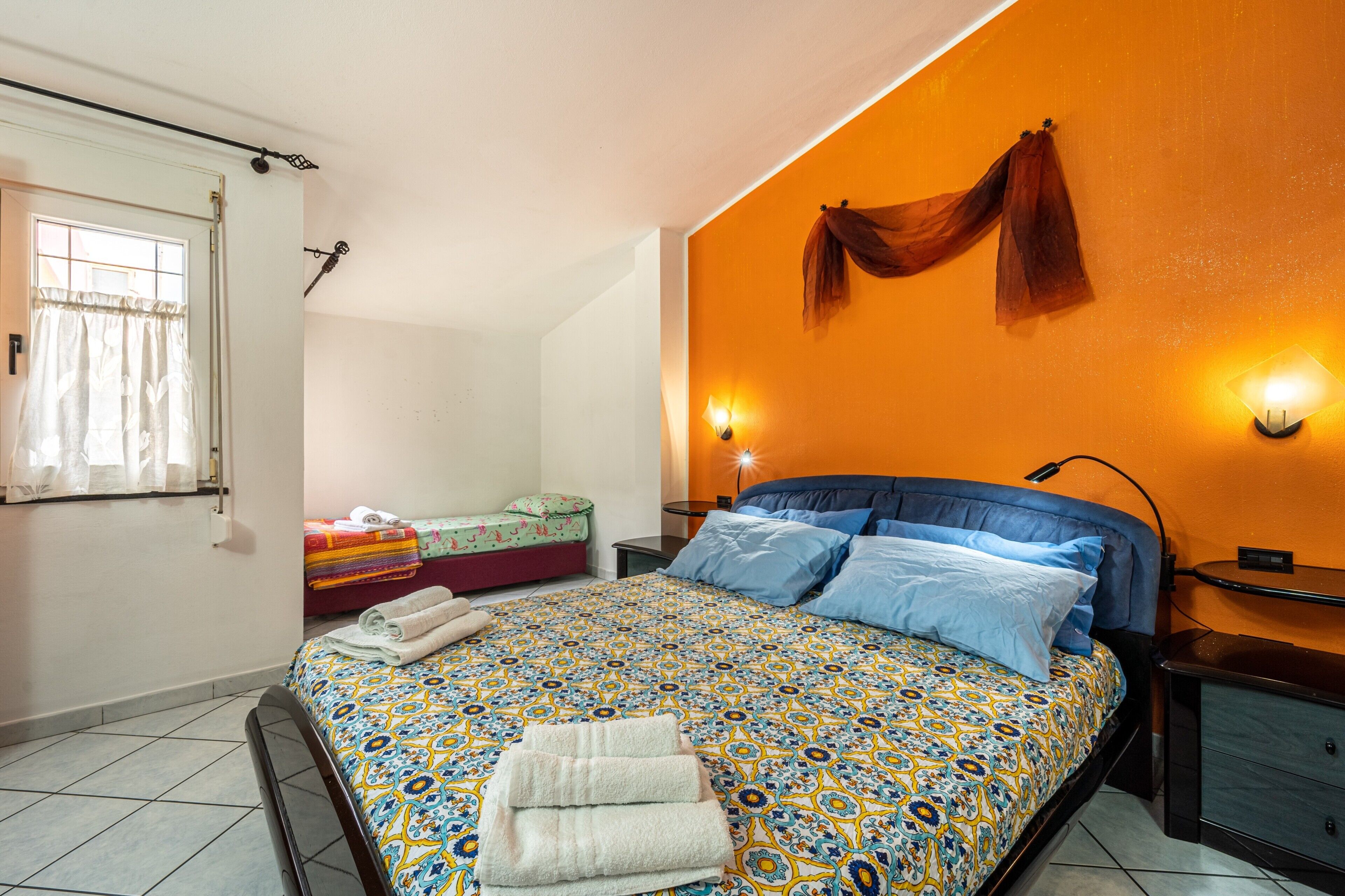 Un dormitorio con una colcha estampada, una ventana con cortinas, una mesita de noche con lámpara y un perchero para toallas en la pared.