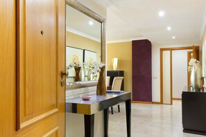 Une chambre d’hôtel avec une porte en bois, une table sur laquelle se trouvent un vase et un bol, un miroir qui reflète la pièce et un mur d’accent de couleur violette.