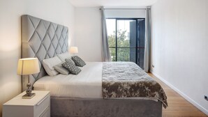 Ett sovrum med en stoppad sänggavel, en säng med ett mönstrat överkast, ett sängbord med en lampa och en utsikt genom ett fönster med gardiner.