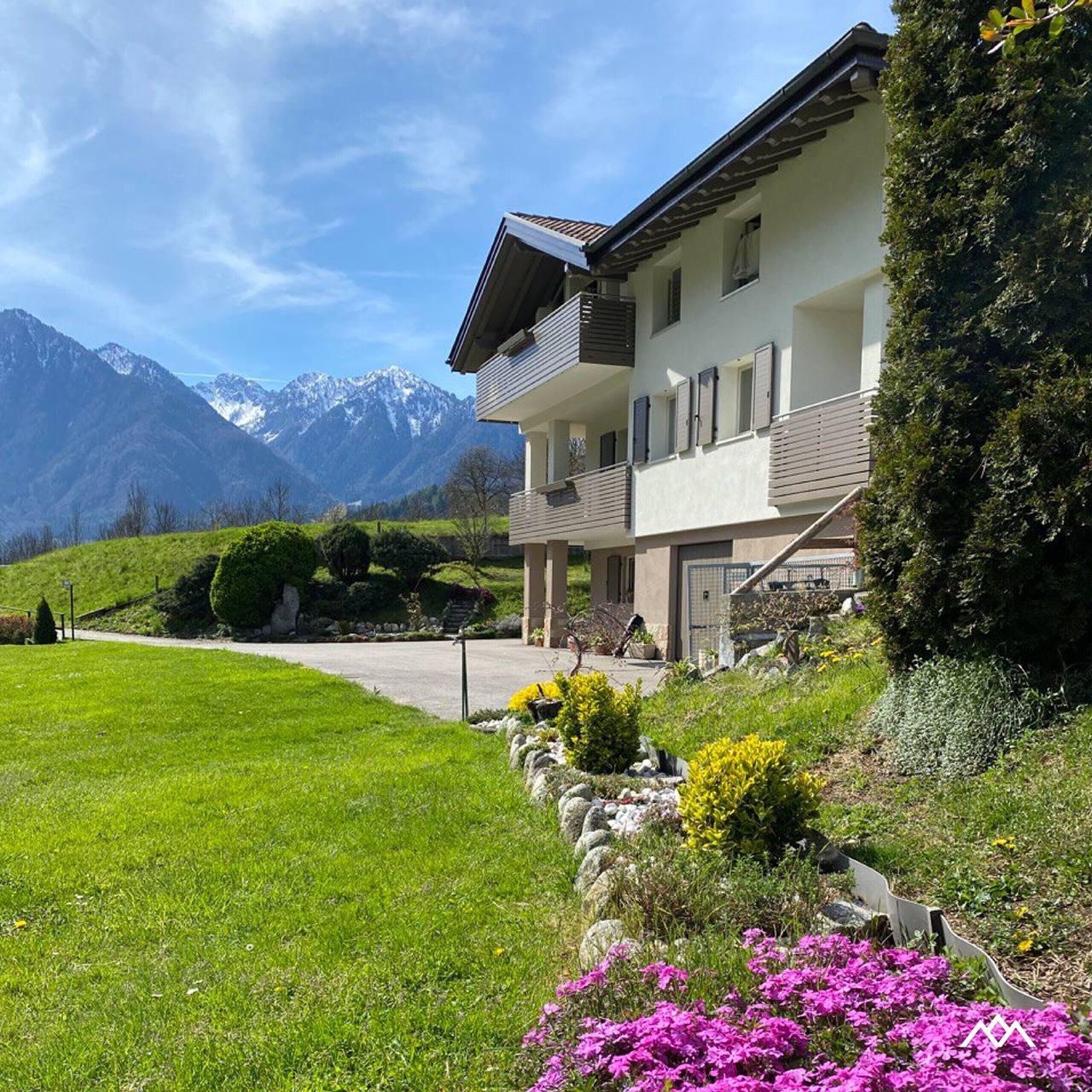 A two-story building with a balcony, a paved driveway, and a mountain backdrop.