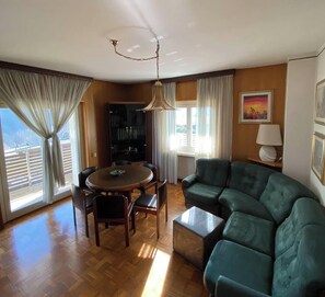 A living room with a green sofa, a dining table with chairs, a chandelier, and a bookshelf.