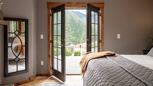 Un dormitorio con una cama grande, piso de madera y vistas a un paisaje montañoso a través de puertas abiertas.