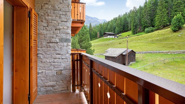 Un balcone in legno con un muro in pietra, vista su un pendio verde e una piccola costruzione in legno in lontananza.