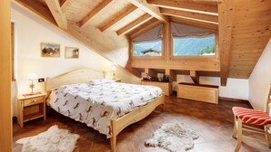 Une chambre avec un plafond en pente, des poutres en bois, un lit recouvert d’une couvre-lit à motifs, une table de chevet avec une lampe, une chaise et une vue sur les montagnes par la fenêtre.