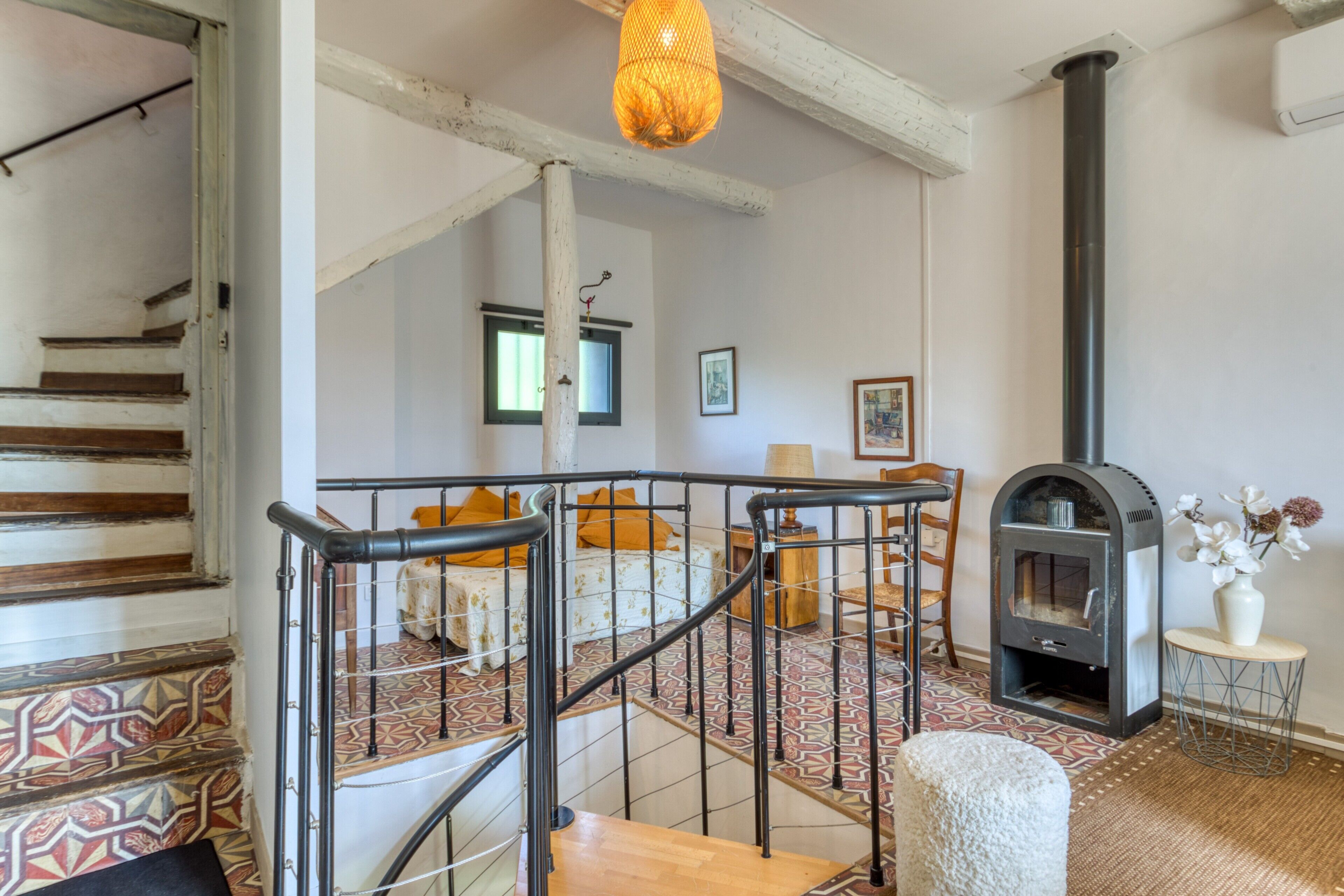A staircase with patterned tiles, a bed with a white and orange bedspread, a wooden chair, a small table with a vase, and a wood-burning stove.