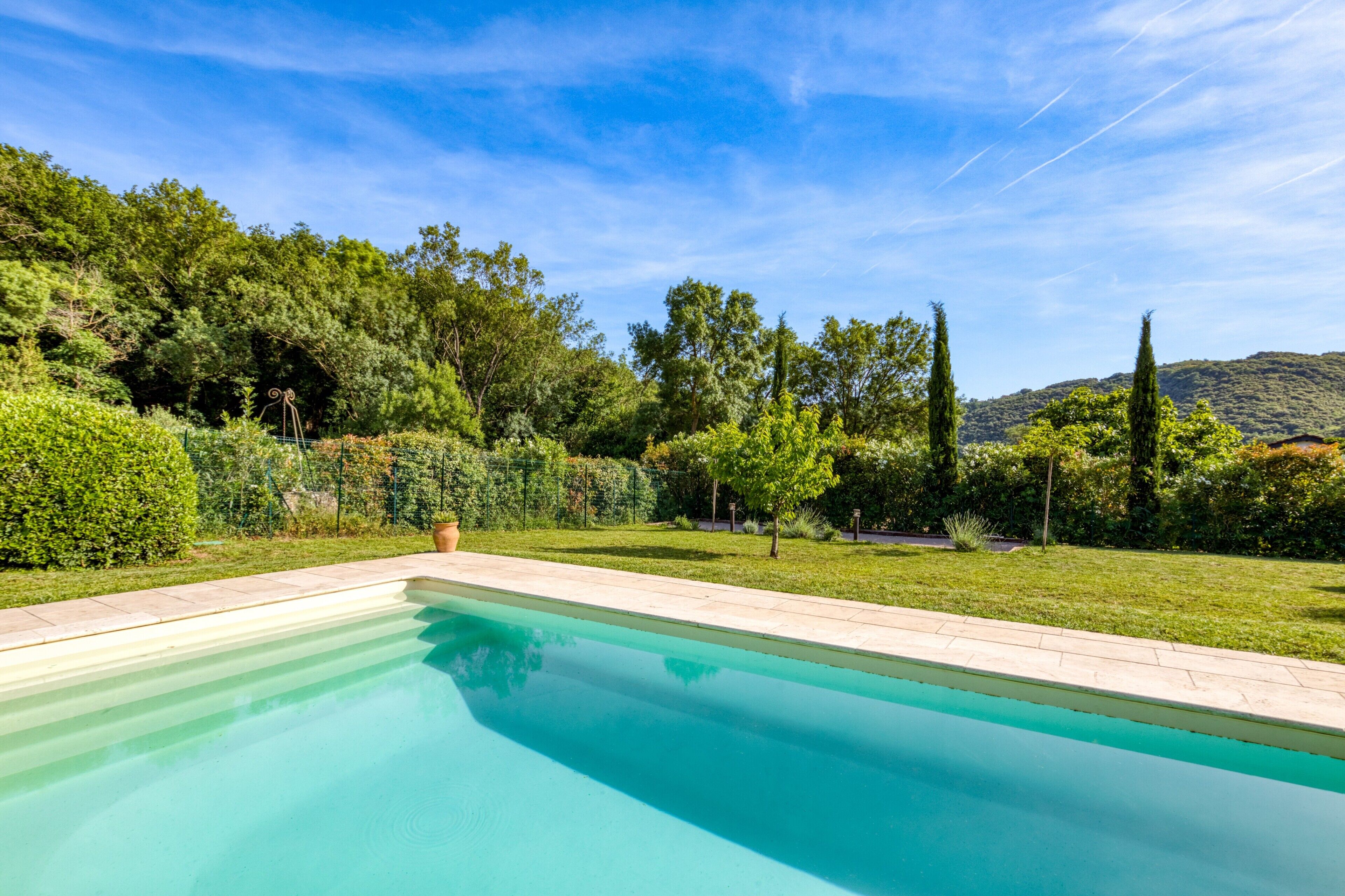 Una piscina rectangular con borde de azulejos, rodeada por un césped cuidado y varias árboles.