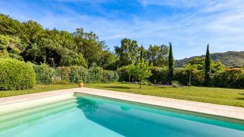 Una piscina rectangular con borde de azulejos, rodeada por un césped cuidado y varias árboles.