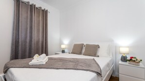 Een net opgemaakt bed met een wit-grijs deken, twee kussens en een opgevouwen handdoek aan de zijkant. Er staan twee nachtkastjes met lampen en een klein decoratief object op een ervan.