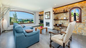 A living room with a sofa, armchair, stone wall, television, fireplace, and a view of the landscape. - Country House 'Es Pujols' with Private Pool, Private Terrace and Wi-Fi (Artá)