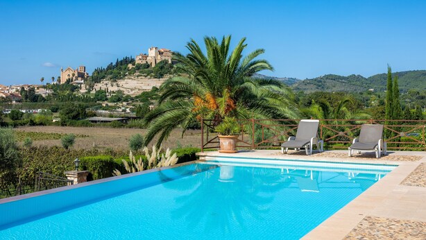 A swimming pool with lounge chairs, a palm tree, and a castle in the background. - Country House 'Es Pujols' with Private Pool, Private Terrace and Wi-Fi (Artá)