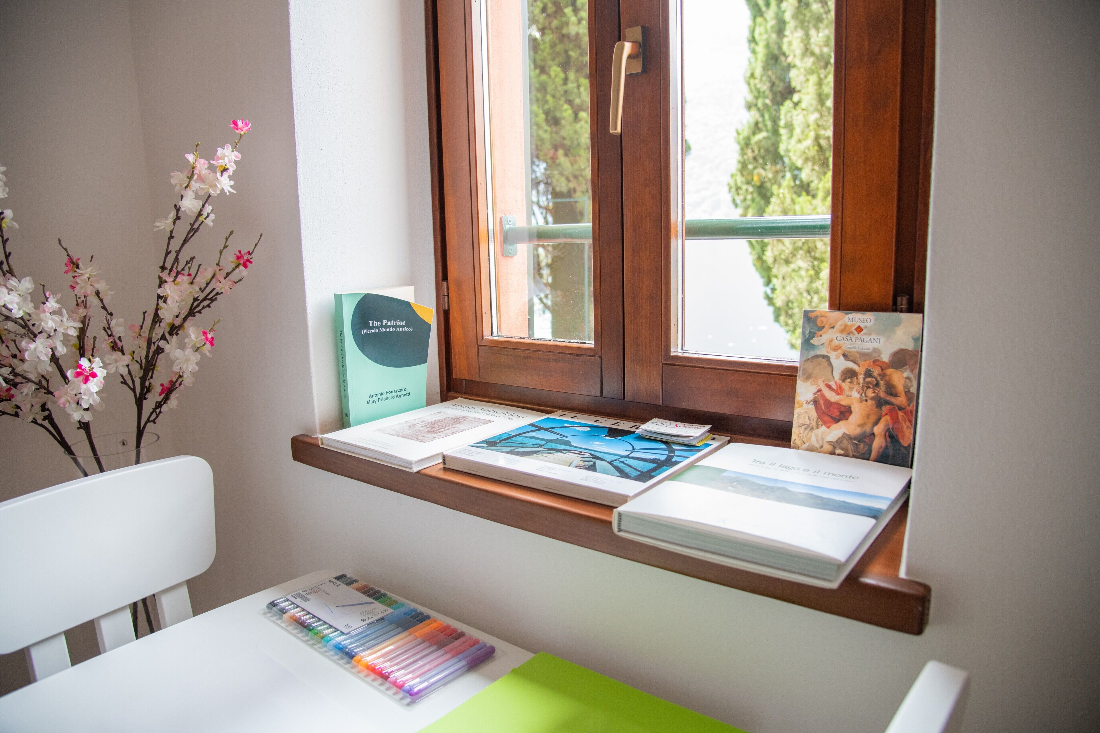 Ein Zimmer mit Fenster, Schreibtisch mit Büchern und Schreibwaren sowie eine Vase mit Blumen.