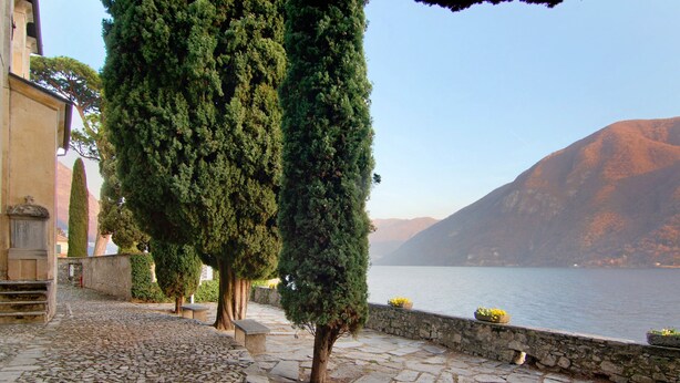A cobblestone pathway, tall trees, a stone wall, and a body of water with mountains in the background. - Apartment 'Fogazzaro Lake View' with Lake View, Wi-Fi and Air Conditioning (Albogasio-Oria Valsolda)