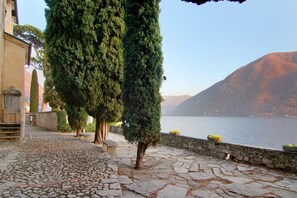 A cobblestone pathway, tall trees, a stone wall, and a body of water with mountains in the background. - Apartment 'Fogazzaro Lake View' with Lake View, Wi-Fi and Air Conditioning (Albogasio-Oria Valsolda)