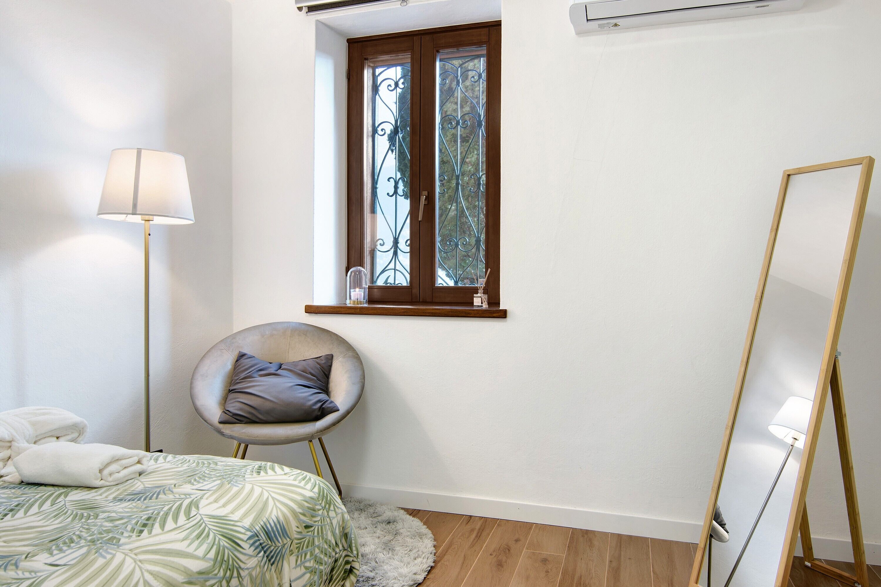 A bedroom with a bed, a chair, a standing lamp, a window with iron bars, and a wooden floor.