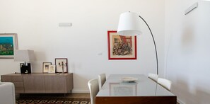Un moderno comedor con una mesa de vidrio, sillas blancas, una alfombra estampada, una lámpara blanca y un cuadro en la pared.