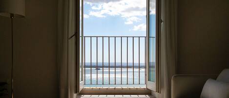 A room with a view of the sea through a window with a metal railing. There is a sofa and a table with a plate on it.