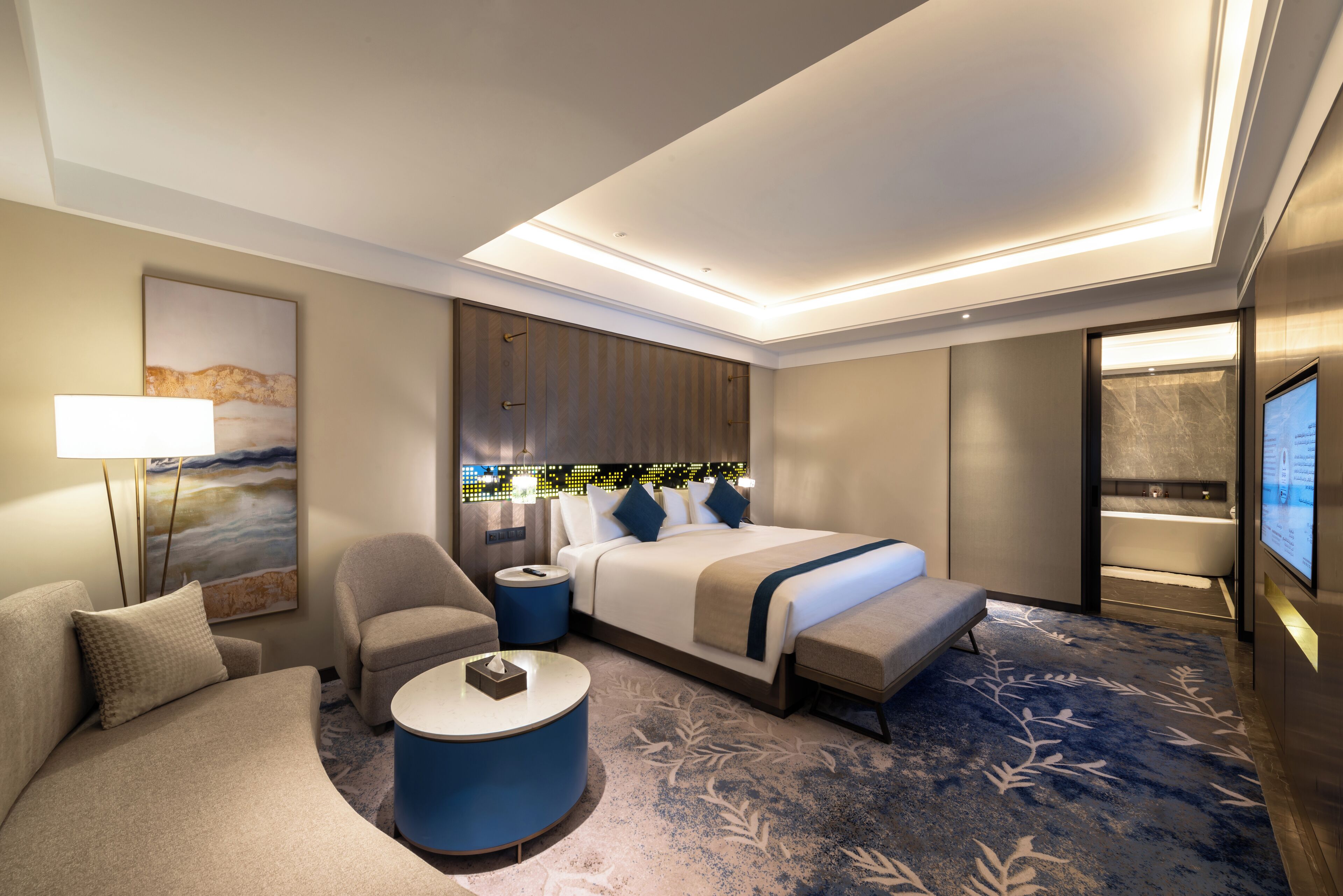 A modern hotel room with a large bed, a sofa, a small round table, and a bathroom visible through an open door.