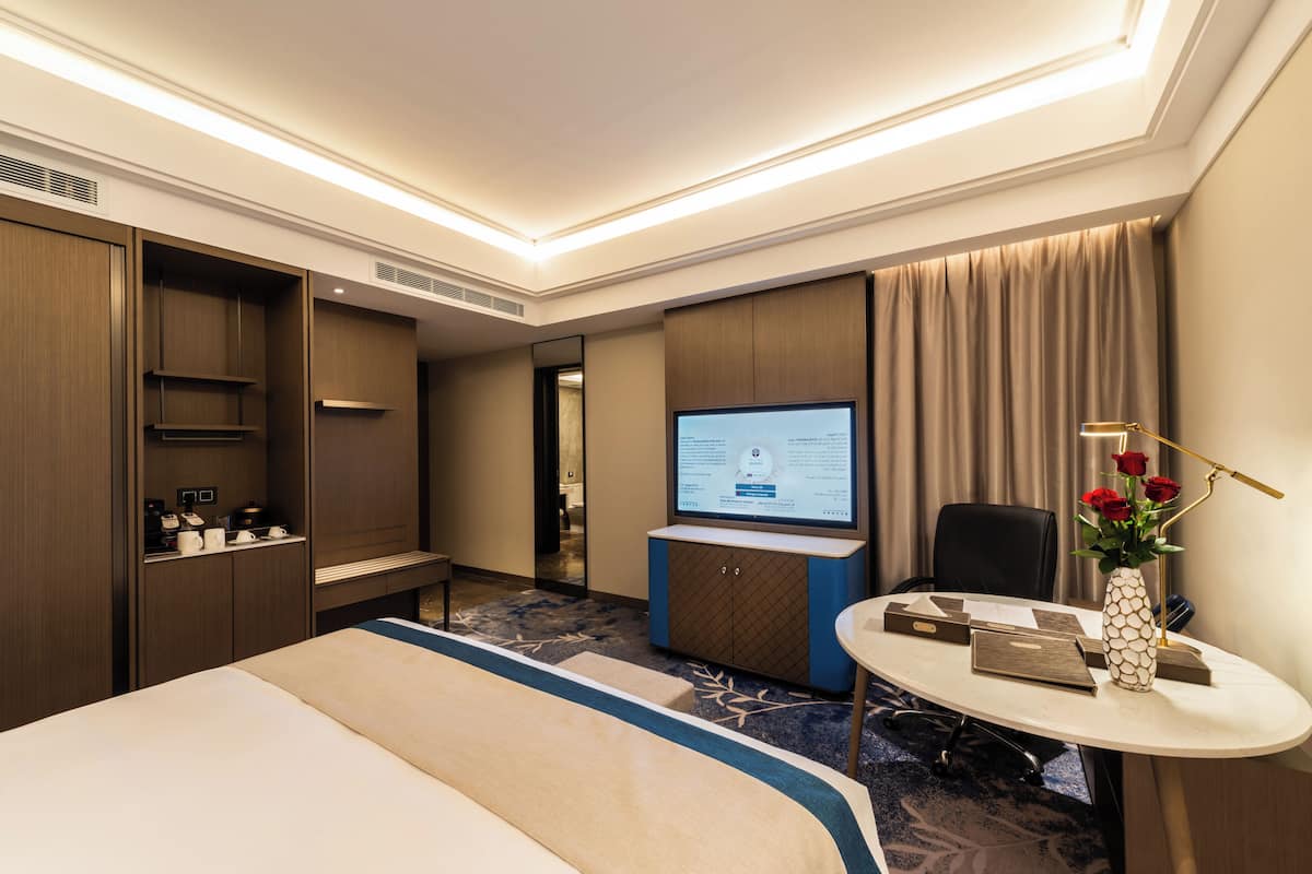 Habitación de hotel con una cama, un escritorio con computadora, una silla, una mesita con un jarrón de flores y un televisor.