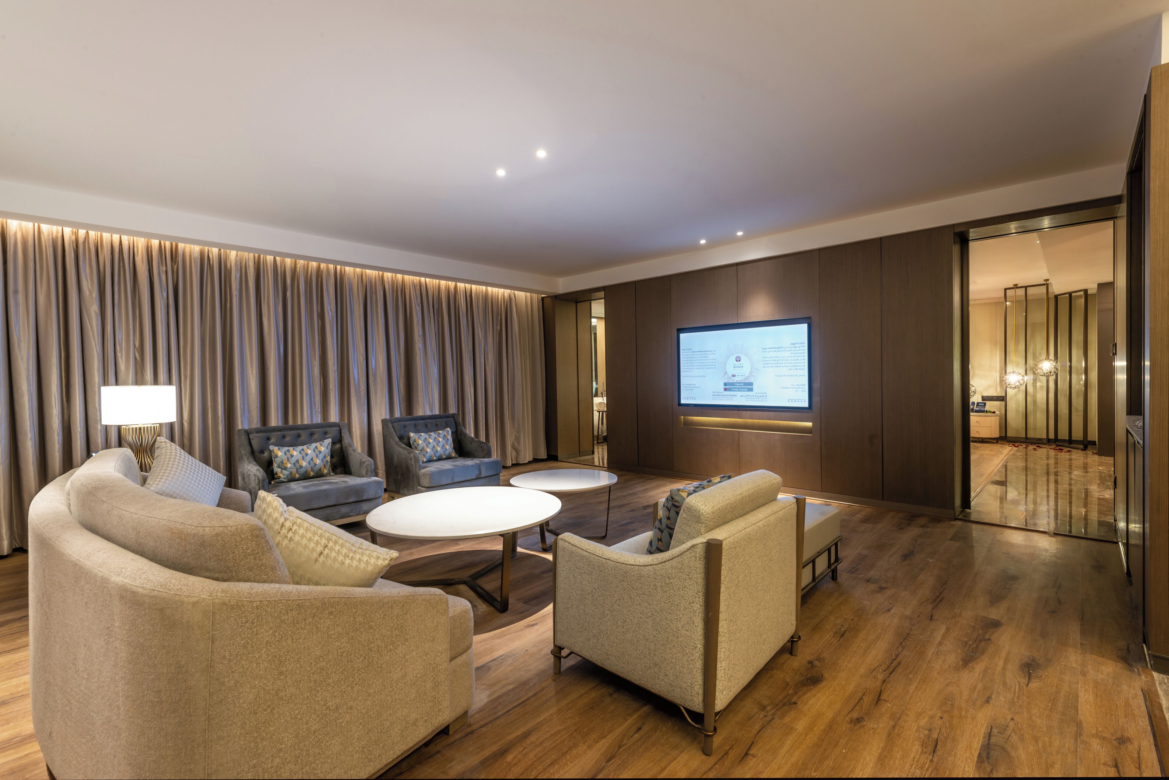 A modern hotel room with a large flat-screen TV, two sofas, a coffee table, and a wooden floor.