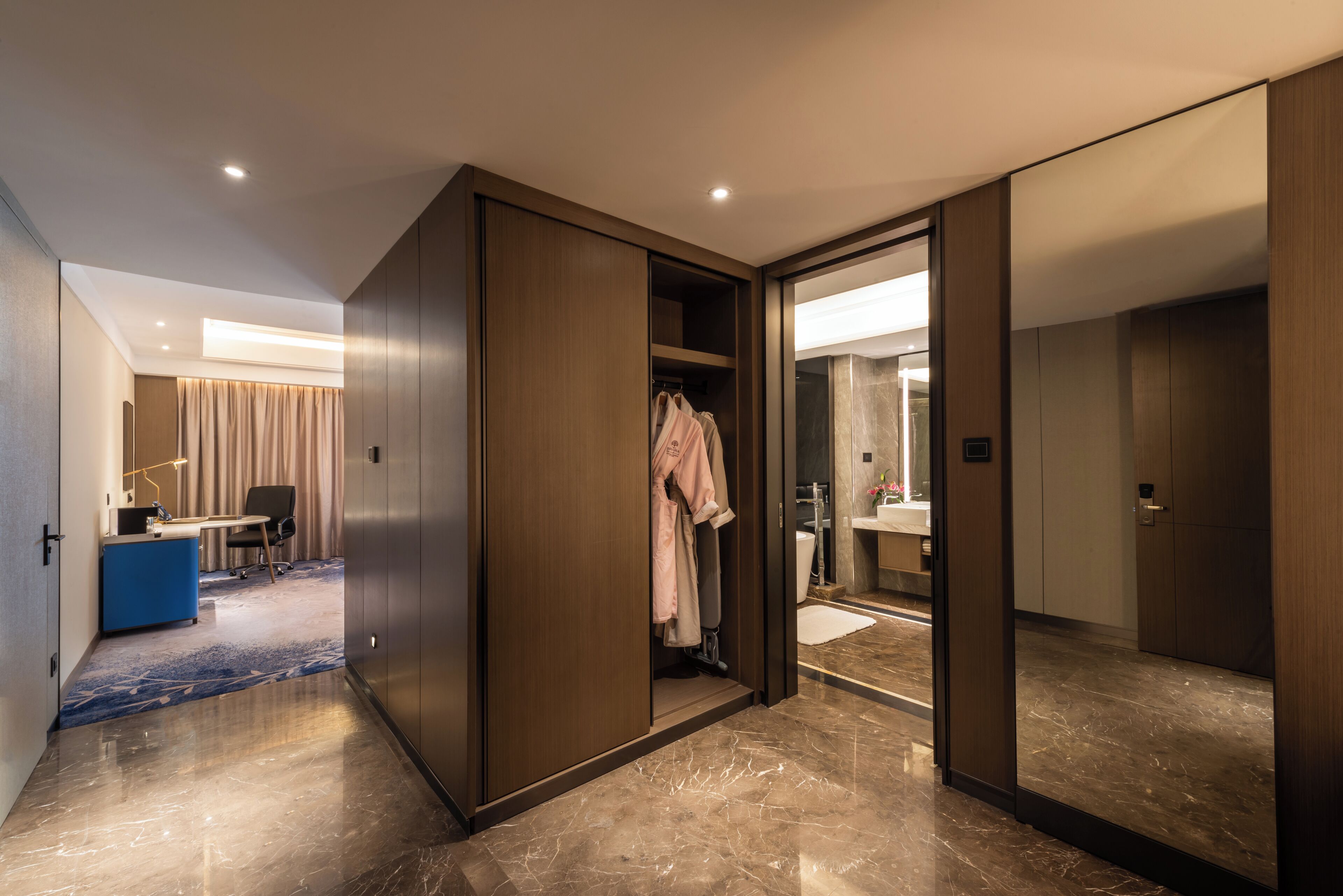 A modern hotel room with a walk-in closet, a desk, and a bathroom with a large mirror.