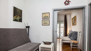 Ein Wohnzimmer mit grauem Sofa, einem kleinen weißen Tisch und einem Sessel. An der Wand hängen zwei gerahmte Bilder, auf einem Tisch steht eine Schale, und ein Durchgang führt in einen anderen Raum mit Bett und Fenster.
