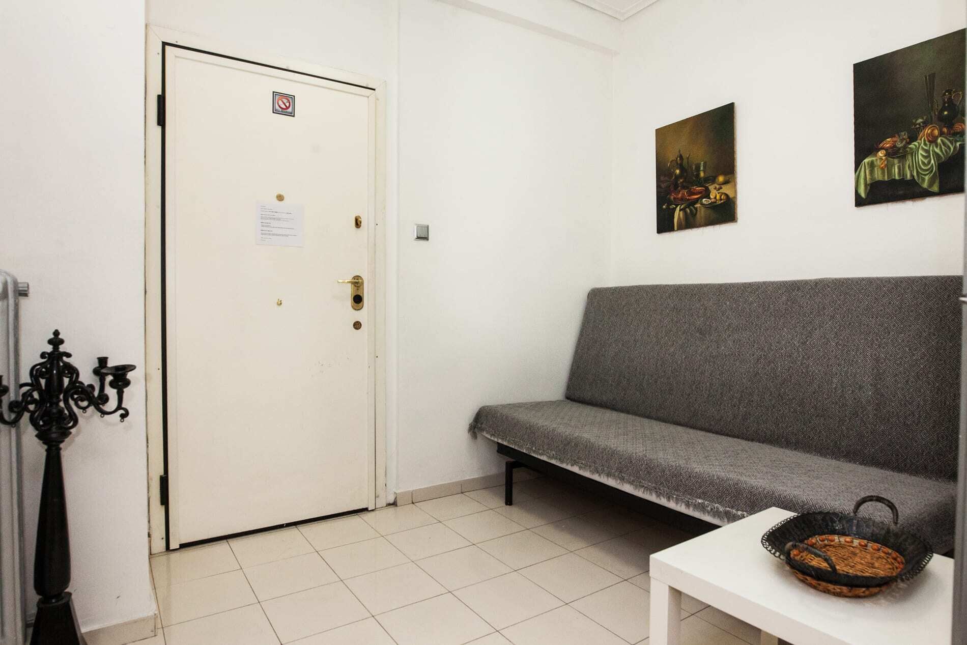A room with a white door, a grey sofa, a white table with a basket, and two paintings on the wall.