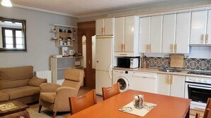 En kompakt opholdsstue med sofa, lænestol, spisebord, tekøkken og indbyggede reoler.