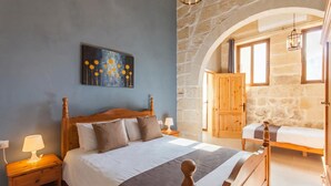 A bedroom with a large bed, a nightstand with a lamp, a painting on the wall, and a stone archway. - Naduri, Beautiful Gozitan Villa + Pool (Nadur)