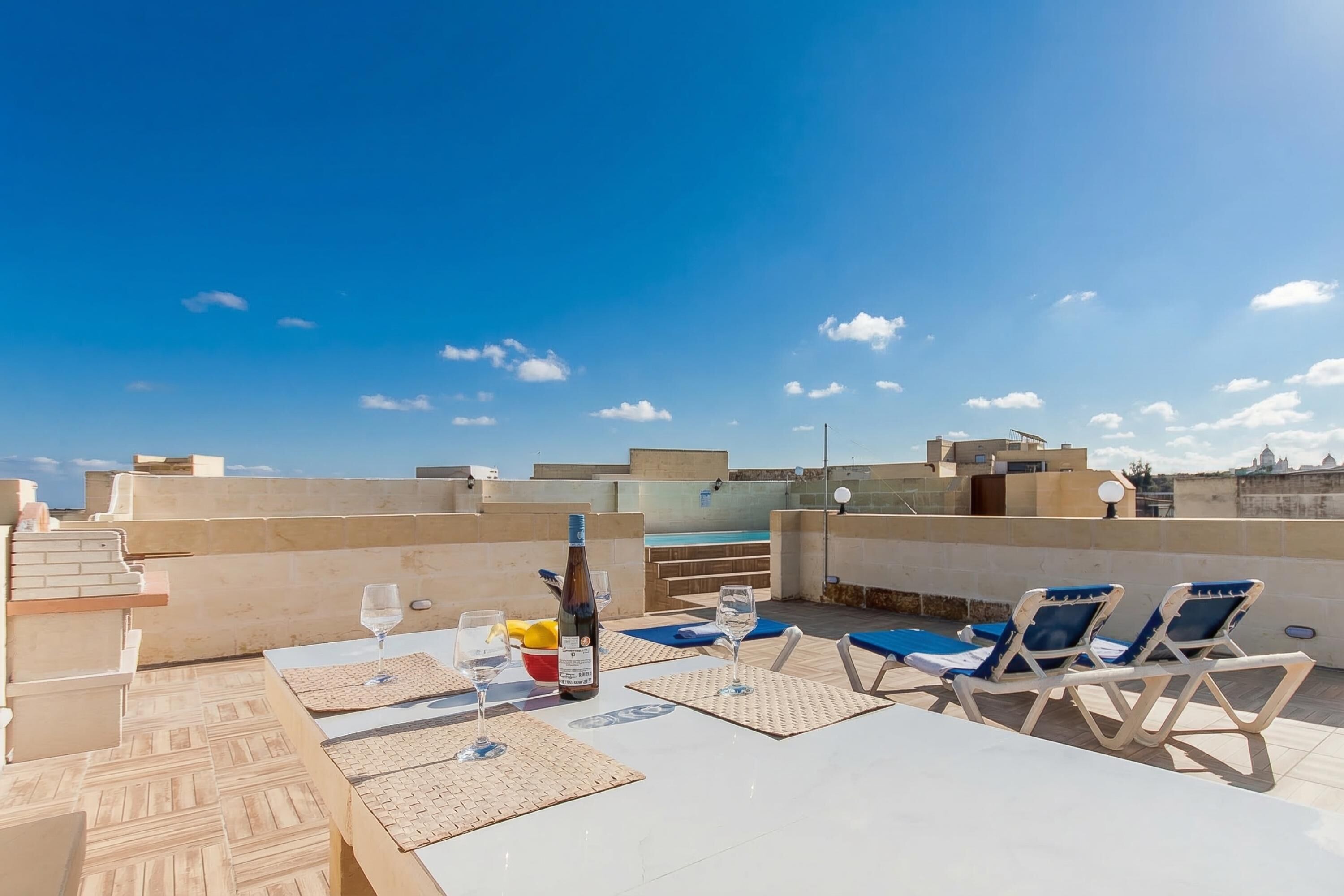A rooftop terrace with lounge chairs, a table with glasses and a bottle, and a pool in the background.