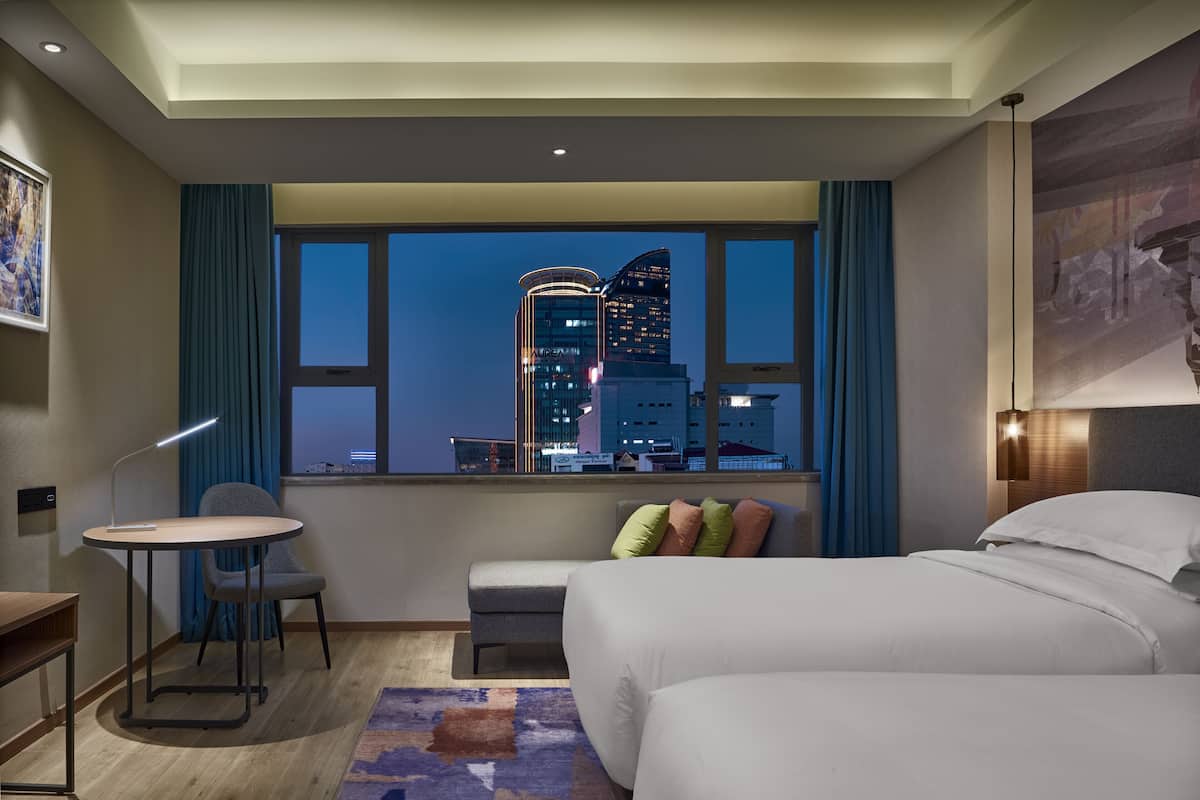 A hotel room with a bed, a desk, a chair, a small table, and a view of a cityscape at night.