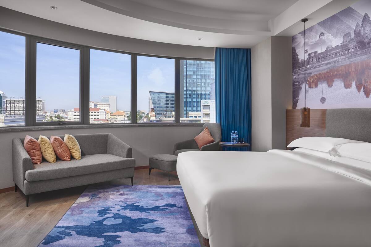 A modern hotel room with a large bed, a sofa, a chair, and a view of the cityscape through large windows.