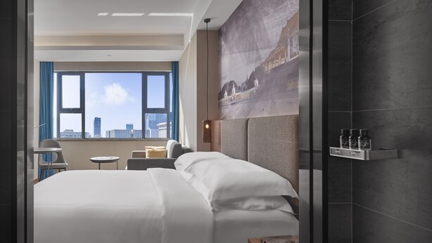 A hotel room with a large bed, a small table, a chair, a view of the city, and a wall-mounted shelf with toiletries. - Aurea Central Hotel (Phnom Penh)