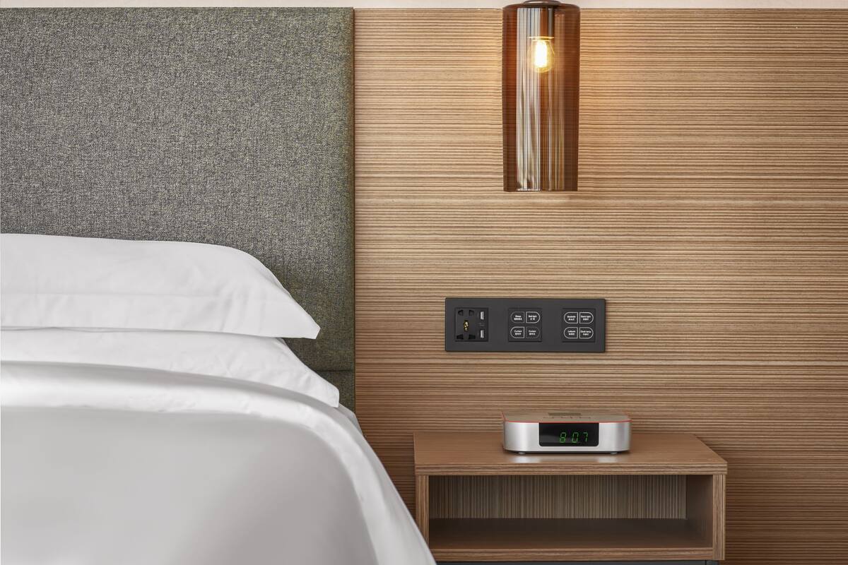 A modern hotel room with a bed, a nightstand with a digital clock, and a wall-mounted light fixture.