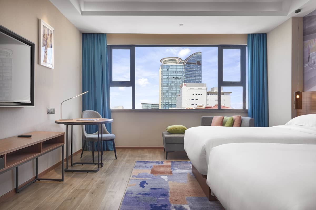 A hotel room with a bed, a desk, a chair, a small table, a nightstand, a lamp, and a view of a cityscape through large windows.