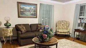 A living room with a brown sofa, a floral armchair, a round coffee table with a flower arrangement, a side table with a lamp, and a framed cityscape painting on the wall.