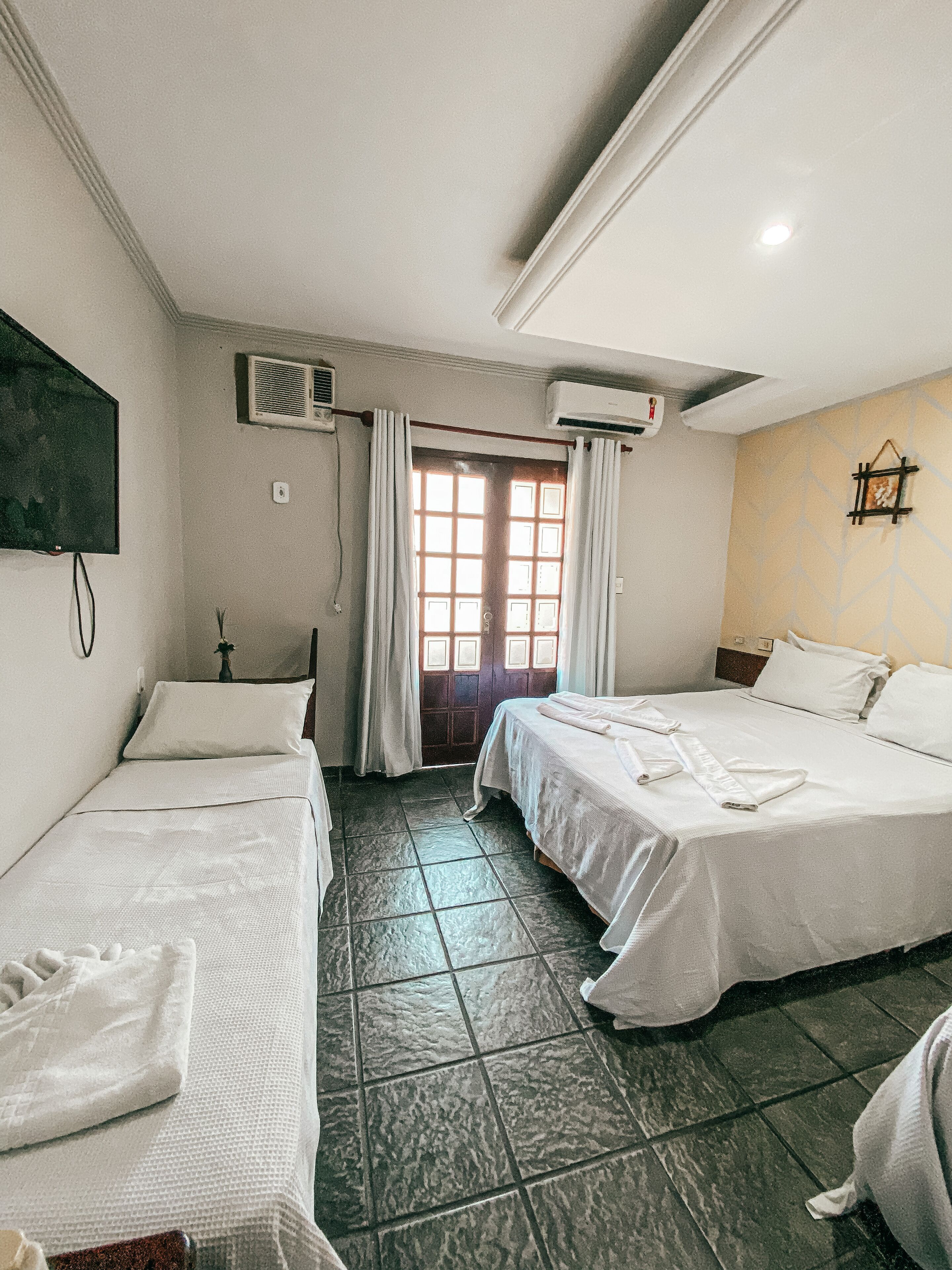 A hotel room with two beds, a flat-screen TV, an air conditioning unit, a door with curtains, and a tiled floor.