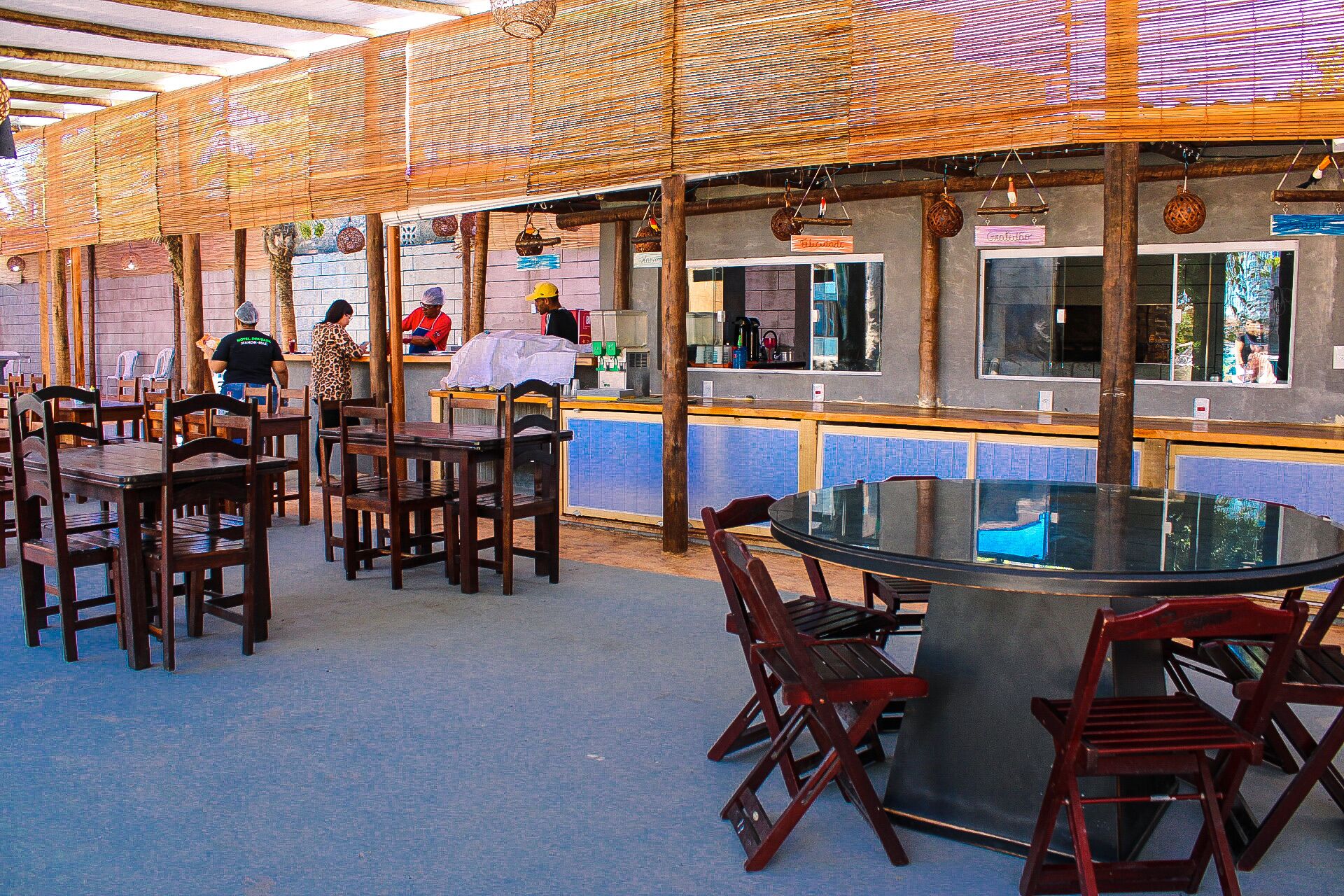 Un restaurante al aire libre con mesas y sillas de madera, una mesa de vidrio central y un techo de bambú.