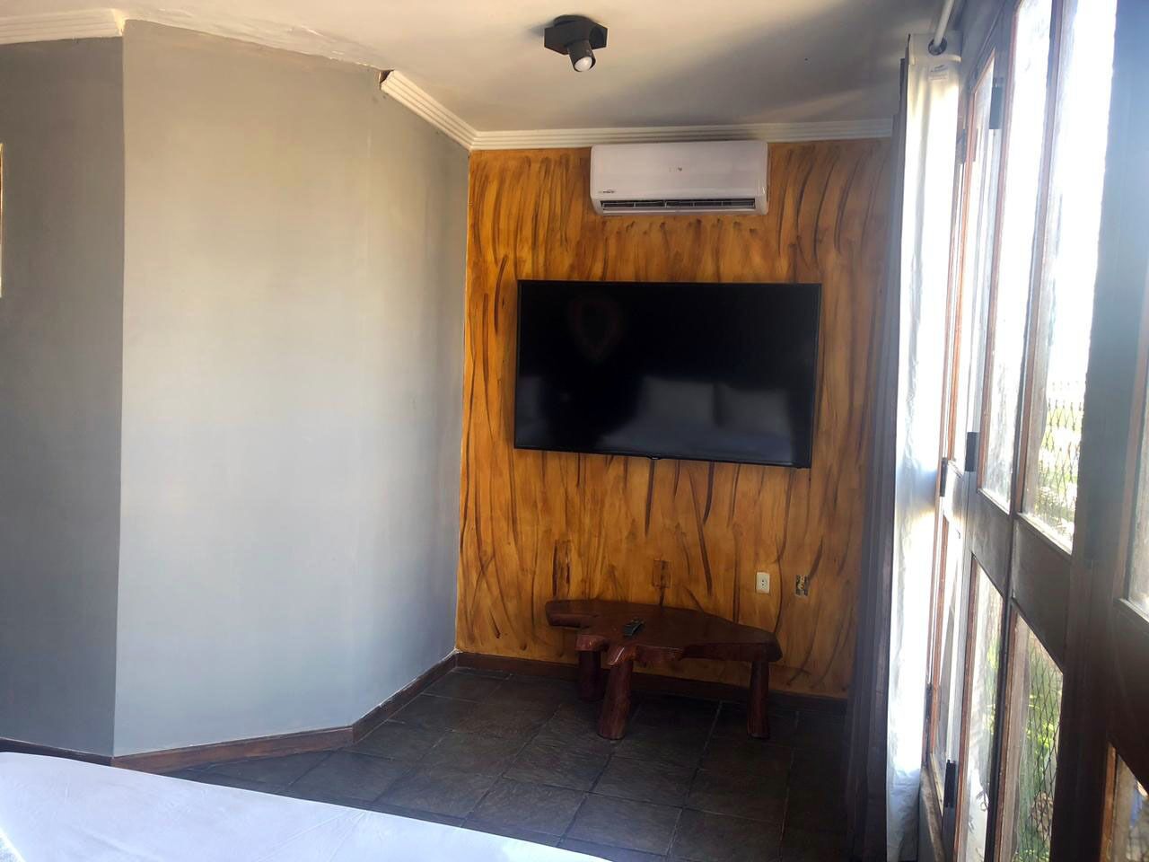 Habitación con televisor de pantalla plana montado en una pared de madera, un pequeño banco de madera y una ventana con cortinas.