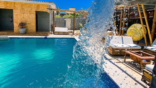 Una piscina azul cristalina con una cascada, rodeada de sillones de madera y una silla tejida amarilla.