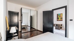 A modern hotel room with a large bed, a desk, and a chair. There is a mirror on one wall and a painting on another.