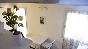 A dining area with a white table and chairs, a vase with flowers, a framed picture on the wall, and a window with curtains.