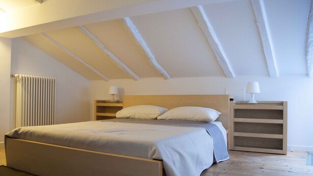 A bedroom with a bed, two bedside tables, a radiator, and a sloped ceiling with exposed beams.