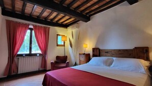 A bedroom with a wooden headboard, a bed with white linens, a chair, a nightstand, and a window with red curtains. - Tenuta Colombaio  (Casole d'Elsa)