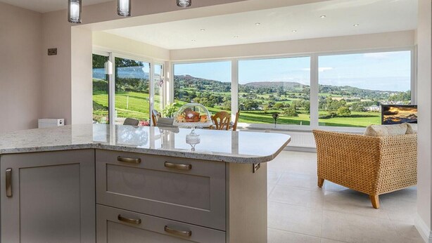 A modern kitchen with a large island, a dining area with a view of a green landscape, and a wicker chair.