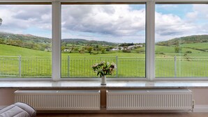 A room with a large window offering a view of a green landscape with houses and hills. There is a white radiator below the window and a small vase with flowers on the windowsill.