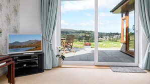 A room with a television, a sliding glass door leading to a patio with a chair and a table, and a view of a landscape with mountains and fields.